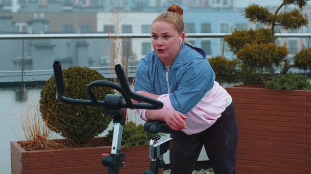 Atletická dívka provádějící aerobní jízdu na koni cvičení na kole stacionární kolo na střeše domu - Fotografie, Obrázek