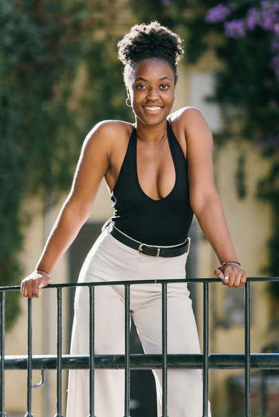 African model leaning on a rail with a black tank top and white trousers while smiling - Foto, afbeelding