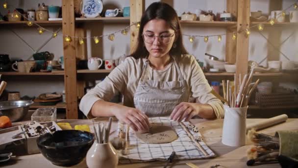 Young woman potter makes a design on the clay plate using a stamp - Felvétel, videó
