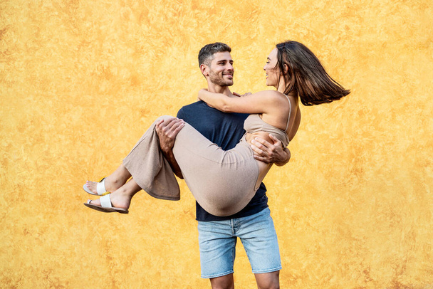 Happy couple on a date. Handsome man carrying his beautiful woman on hands. Yellow wall. A lot of copy space. Real people emotions.  - Foto, Imagem