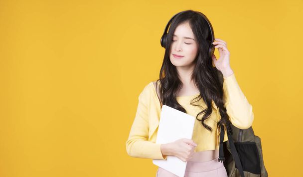 Радостная студентка азиатского университета держит планшетный компьютер урок прослушивания онлайн с беспроводным наушником на изолированном желтом фоне. Подростковая азиатка думает, что-то расслабиться позитивность. - Фото, изображение