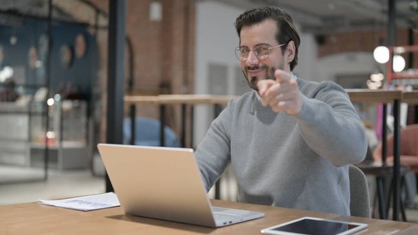 Young Man Pointing at Camera while using Laptop in Office - Foto, afbeelding