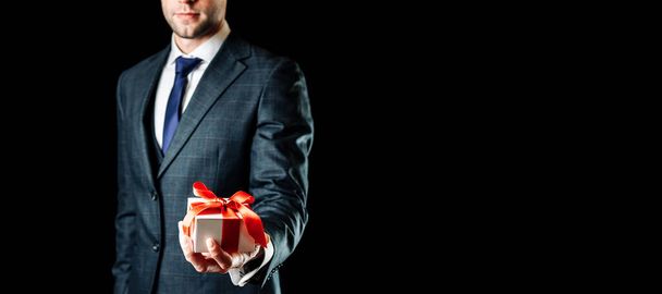 ギフトボックスを持ってる。黒を基調とした赤いリボンで存在感のあるサプライズギフトボックスを持つ幸せな若いビジネスマン。誕生日、バレンタインデー、クリスマス、新年のプレゼント. - 写真・画像