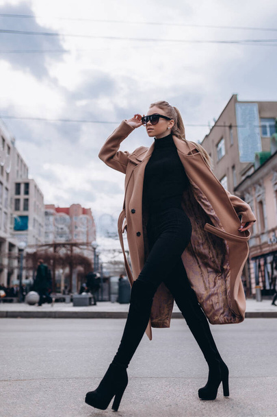 Ein junges schönes Mädchen in beigem Mantel und Sonnenbrille geht durch die Stadt. Bekleidungswerbung - Foto, Bild