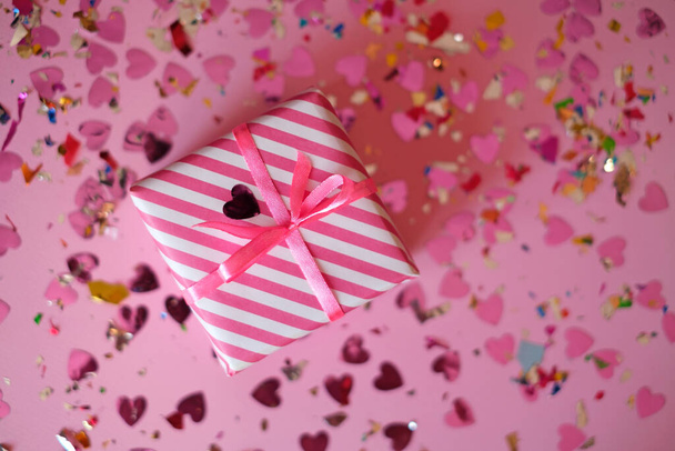 Present in a striped paper wrapper with a pink ribbon on a background with a multicolored shiny confetti of hearts for Valentine's Day. Gift box for the holiday. - Foto, Imagem