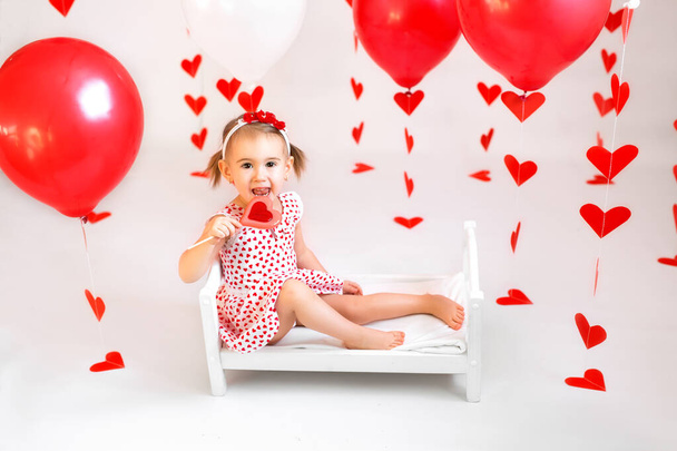 Girl with ponytails is sitting on a small crib with a large red heart-shaped lollipop. Decorations of balloons and garlands with hearts for Valentine's Day - Zdjęcie, obraz