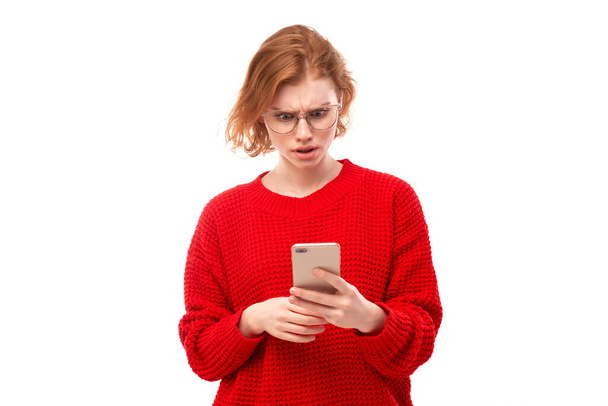 Pensive thinking redhead girl looks into smartphone screen with upset face in glasses and red clothes isolated on white studio background - Valokuva, kuva