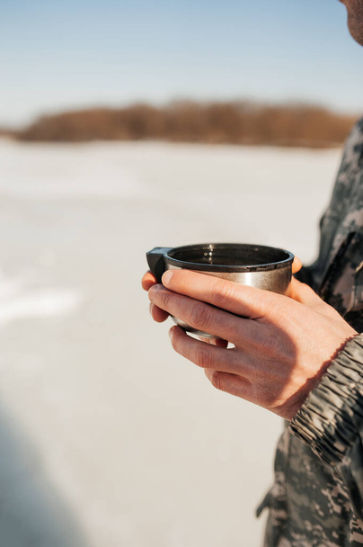 Rude men's hands with dry skin hold a mug of hot tea against the backdrop of military clothing and a winter snowy landscape, vertical travel content, selective focus - 写真・画像