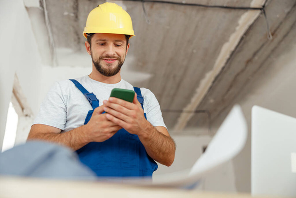 Focused repairman in workwear and hardhat smiling while using smartphone, standing indoors during renovation work - Valokuva, kuva