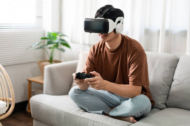 Technology Concept Una persona che indossa un auricolare di realtà virtuale e tiene in mano un gioco di console nero mentre è seduta sul divano. - Foto, immagini