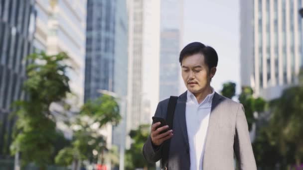 νεαρός ασιάτης επιχειρηματίας με τα πόδια στην κεντρική επιχειρηματική περιοχή κοιτάζοντας κινητό τηλέφωνο στη σύγχρονη πόλη - Πλάνα, βίντεο