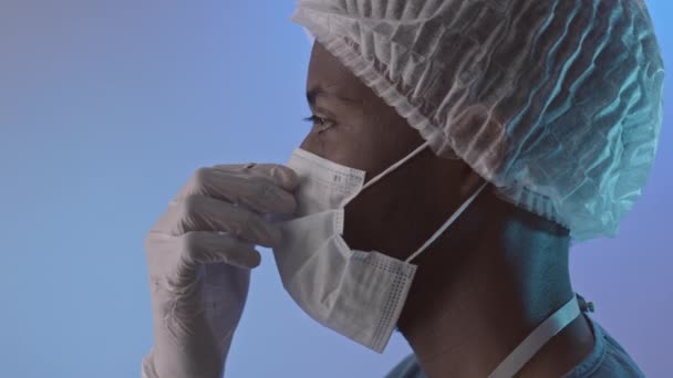 Slowmo side-view PAN close up of young African-American male surgeon taking off face mask and looking ahead standing on light blue background in studio - Video