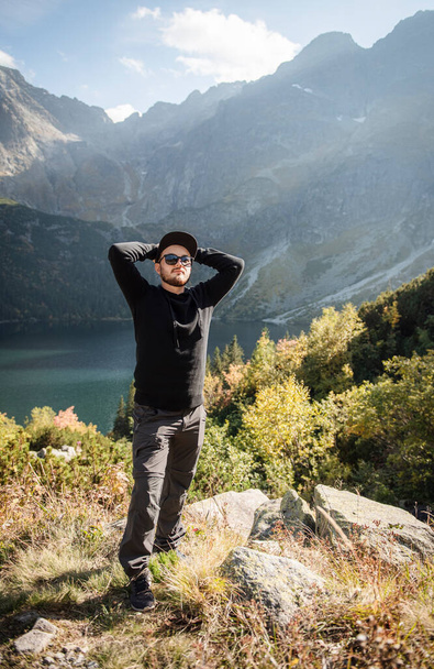 Young tourist man in a cap with hands up on the top of the mountains admires the nature - 写真・画像