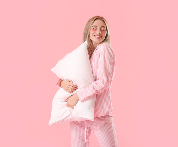 Mujer joven y bonita con parches debajo de los ojos que abrazan la almohada sobre el fondo rosa - Foto, imagen
