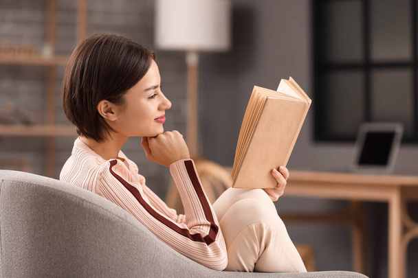 Beautiful young woman reading book in armchair at home - Foto, Imagem