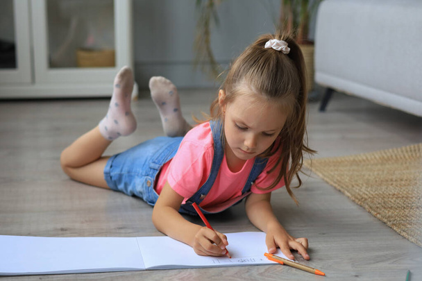 Smilling happy girl lying on warm floor with a toy elephant near to her enjoying creative activity, drawing pencils coloring pictures in albums - Valokuva, kuva