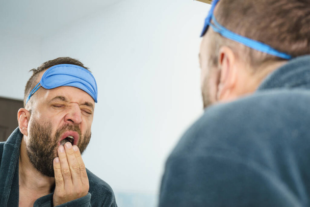 Funny adult man wearing eyemask on forhead having troubles with waking up. Standing in front of mirror looking at his face, yawning, being tired and sleepy. - Foto, imagen