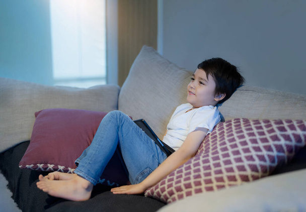 Happy boy with smiling face sitting on sofa watching cartoon, School Kid sitting alone on couch holding tablet and watching TV, Portrait Child relaxing in living room after back from school. - Photo, Image