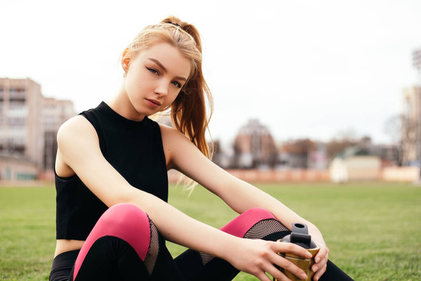 A girl sits on football field, looks at the camera and smiles with metal shaker bottle. Sporty woman in sportswear on the green lawn or grass with copy space. Health care concept - Foto, immagini