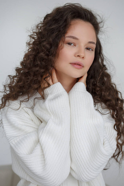 Delicate portrait of a beautiful girl with long curly hair in a cozy white sweater inside. - Valokuva, kuva