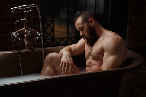 Athletic bearded guy relaxing in a stylish bath in a dark loft premises. - Фото, зображення