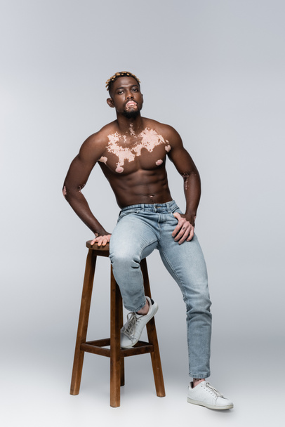 shirtless african american man with vitiligo and stylish hairstyle posing on high stool on grey - Foto, imagen