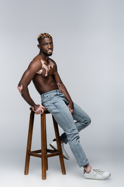 african american man with vitiligo posing shirtless near high stool on grey - 写真・画像