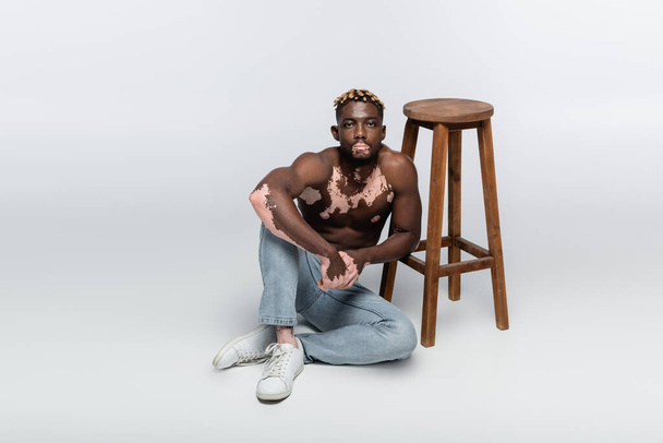 muscular african american man with vitiligo skin and stylish hairstyle sitting shirtless near wooden stool on grey - 写真・画像