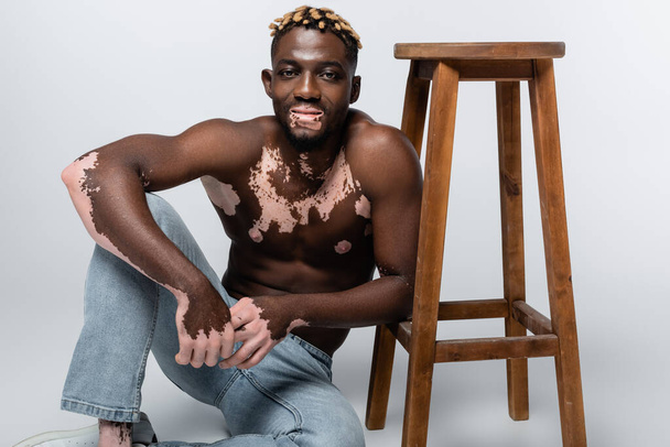 smiling african american man with vitiligo skin and muscular torso sitting near wooden stool on grey - Photo, image