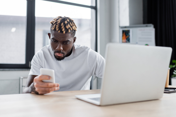 young african american man with vitiligo looking at mobile phone near blurred laptop in office - Foto, Bild