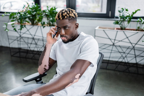 young african american man with vitiligo and trendy hairstyle talking on smartphone in office - Foto, afbeelding