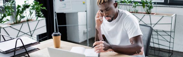 hombre afroamericano con vitiligo celebración de pluma cerca de portátil mientras habla en el teléfono inteligente en la oficina, pancarta - Foto, Imagen