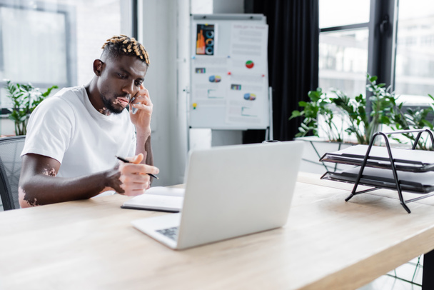 african american man with vitiligo holding pen near notebook and laptop while talking on smartphone in office - Photo, image