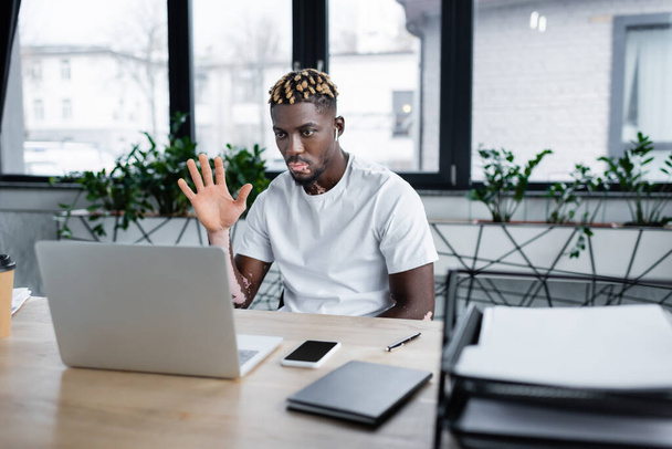 trendy african american man with vitiligo waving hand during video call on laptop in office - Fotó, kép