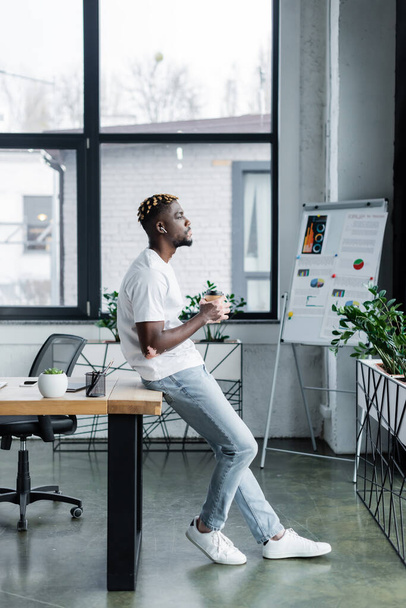 side view of african american man with vitiligo standing with coffee to go near flip chart in office - Foto, Bild