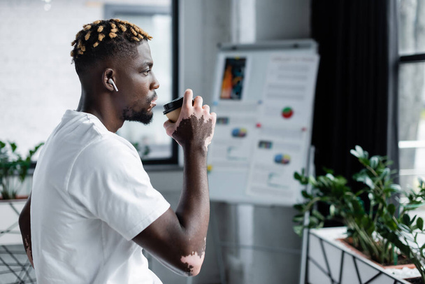side view of african american man with vitiligo drinking coffee and listening music in earphone near blurred flip chart - Foto, Imagen