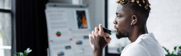 african american man with vitiligo skin and trendy hairstyle drinking coffee to go in office, banner - Foto, afbeelding