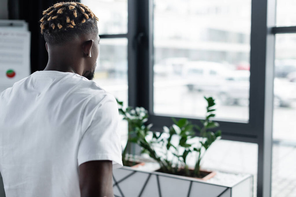 back view of african american man with trendy hairstyle listening music in earphone in office - Foto, afbeelding