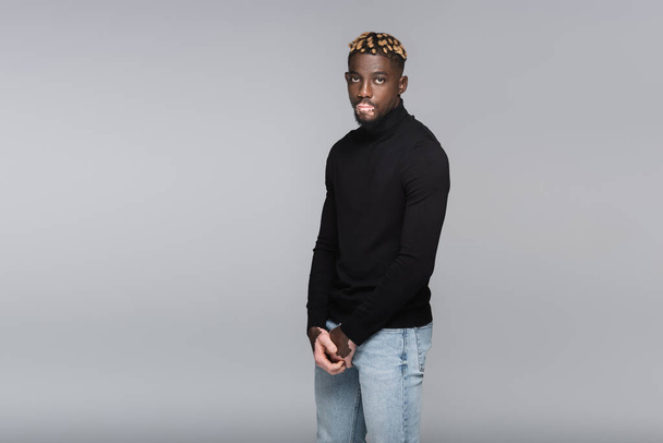 trendy african american man with vitiligo standing in black turtleneck and jeans isolated on grey - Foto, Imagen