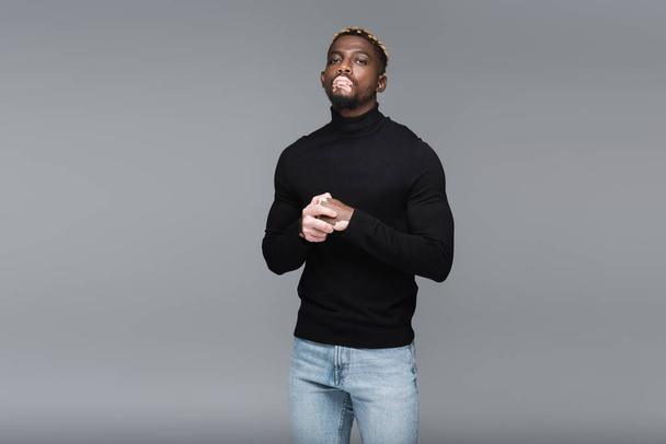 stylish african american man with vitiligo, wearing jeans and black turtleneck, looking at camera isolated on grey - Fotografie, Obrázek