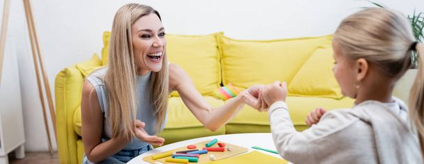 Mother pointing at plasticine near blurred daughter at home, banner  - Фото, изображение