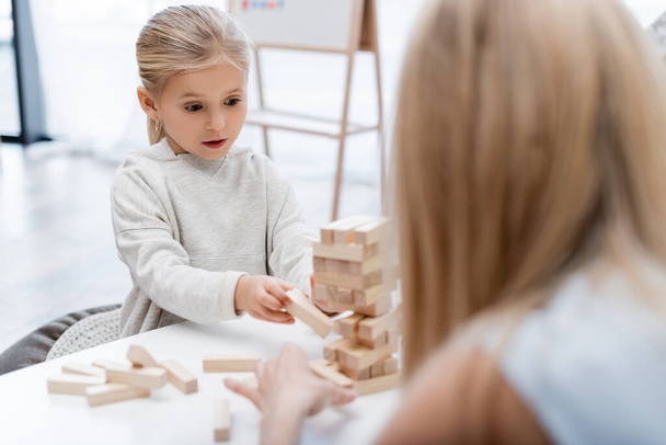 Girl holding wooden block while playing with blurred mother at home  - Foto, Imagen