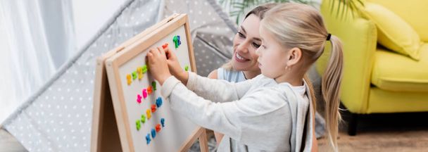 Woman looking at magnetic easel near kid at home, banner  - Foto, imagen