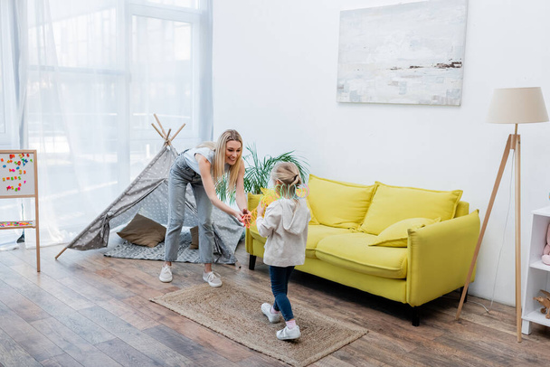 Smiling mother holding toy near daughter, teepee and magnetic easel at home  - Photo, image