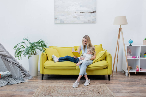 Позитивна жінка читає книгу біля дочки на дивані вдома
  - Фото, зображення