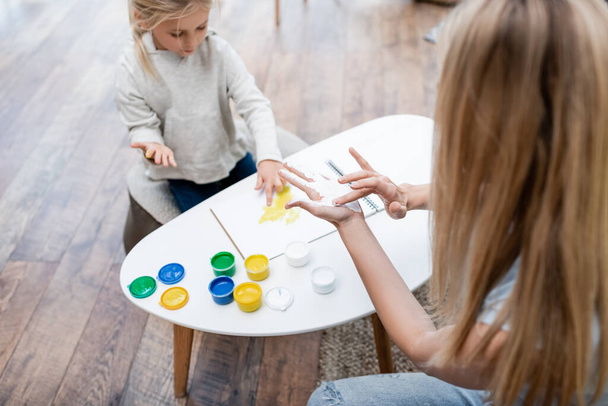 Blurred woman applying paint on hand near kid and sketchbook at home  - Fotó, kép