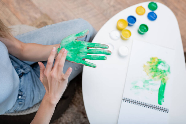 Κάτοψη της γυναίκας που εφαρμόζει χρώμα στο χέρι κοντά σε θολή μπλοκ σχεδίασης στο σπίτι  - Φωτογραφία, εικόνα