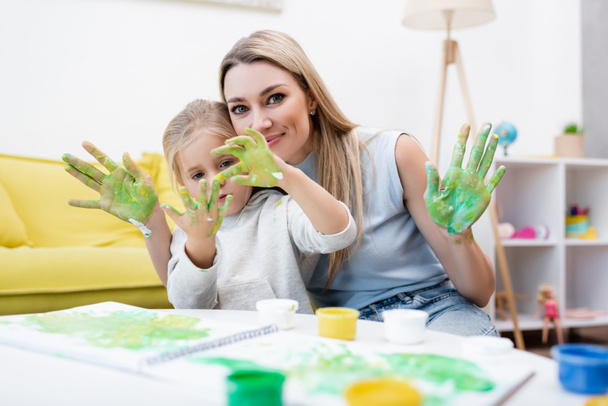 Woman looking at camera near daughter looking at hands in paint at home  - Zdjęcie, obraz