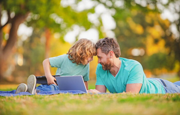 Happy father with son use laptop. Dad and school boy child looking screen and tablet, watching video lesson, sitting on grass. Family weekend. Video call of a grandson to grandparents. - Φωτογραφία, εικόνα