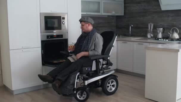 Largo tiro de hombre caucásico en silla de ruedas en su cocina moderna en casa, degustando y disfrutando de galletas calientes frescas del horno durante el día - Imágenes, Vídeo
